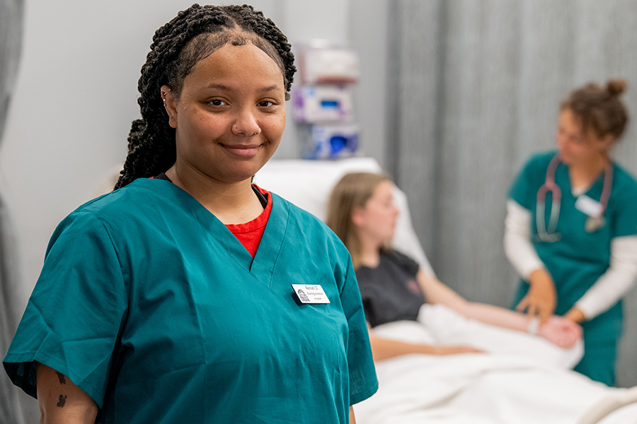 Nursing student in scrubs stands in a simulation lab while another student attends to a patient in a hospital bed