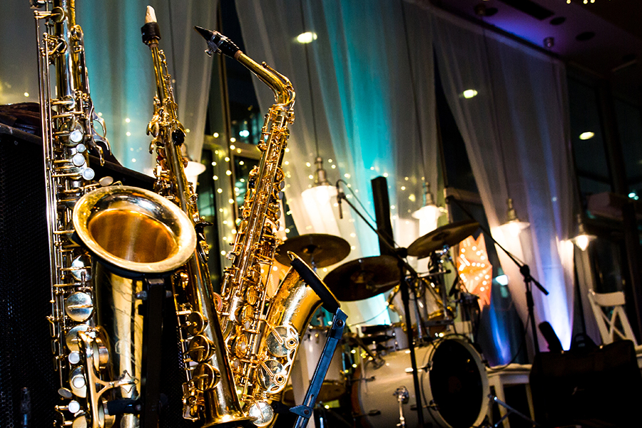 Close-up of saxophones and a drum set on a stage lit with blue and warm lighting.