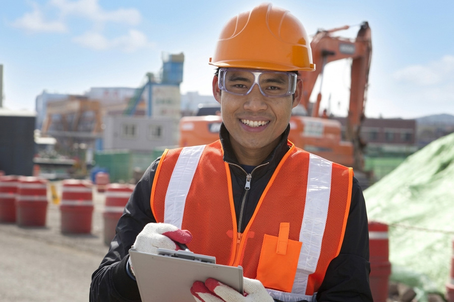 Worker in an orange hard hat and safety vest holds a clipboard on an outdoor job site